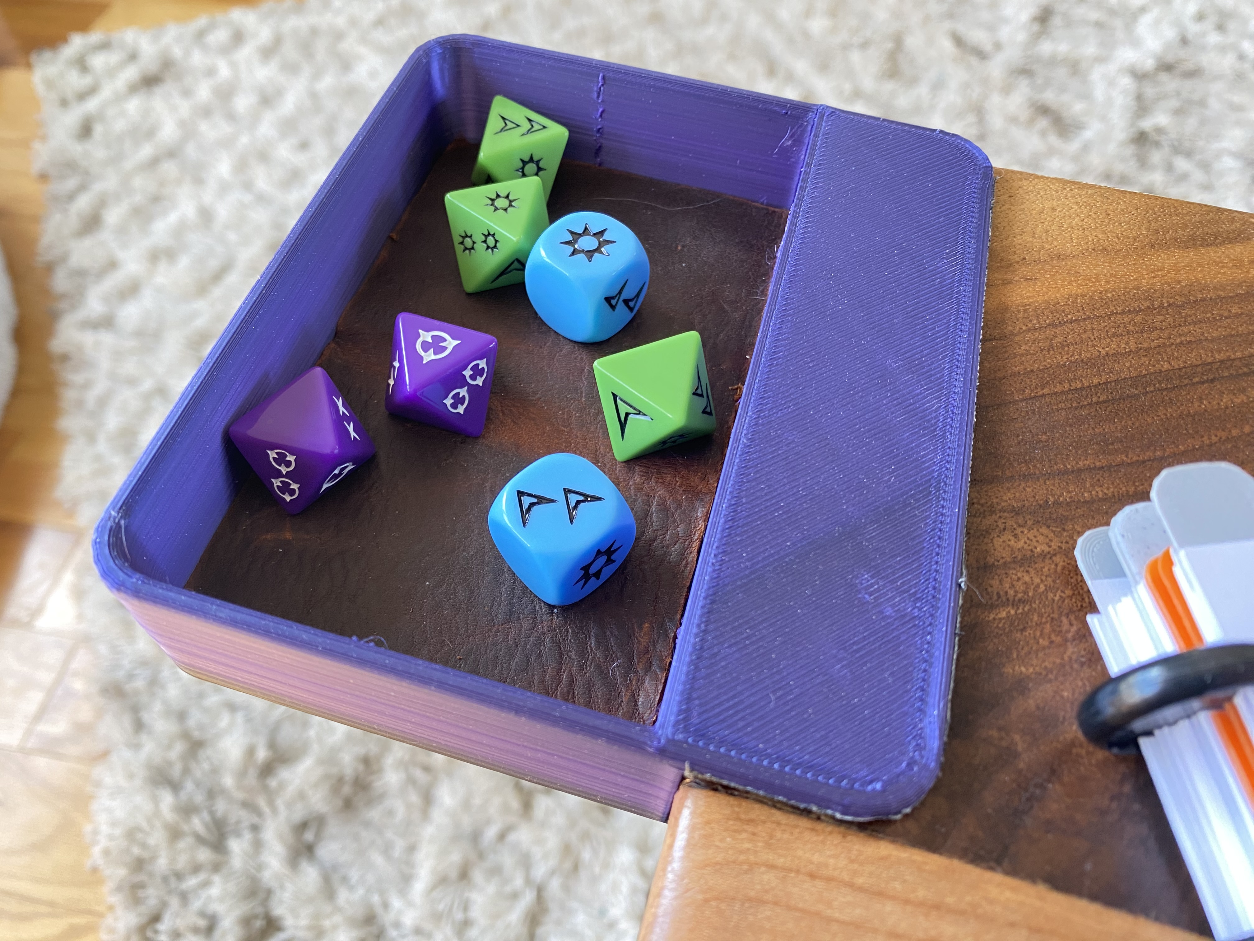 a close-up photo of a 3d printed dice tray holding on to a piece of wood with a notebook on it. in the tray are 7 Genesys dice. The result of the rolled dice is 2 successes and 3 advantages. There is a pieces of brown leather in the bottom of the tray.