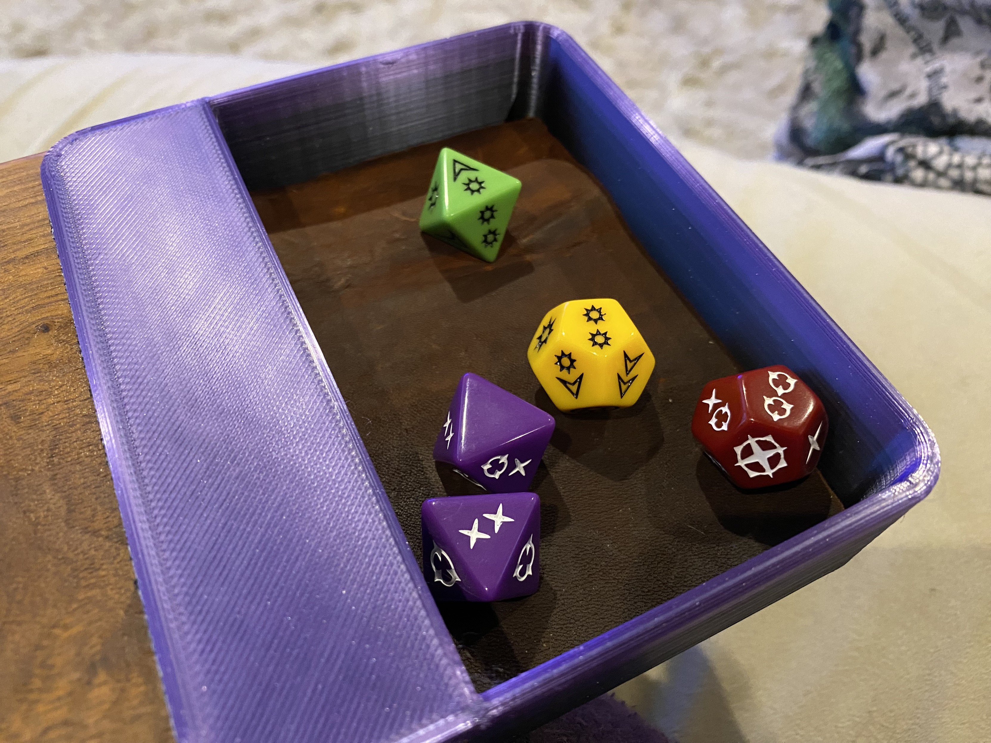 a close-up of small purple dice tray with a lip that is slid over a wooden lap desk. The bottom of the dice tray is lined with leather and has 5 colorful dice in it from the Genesys System Role Playing Game