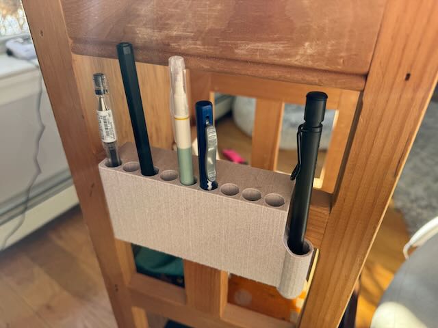 an off-white penn holder with slots for pens that are half-filed. It's slid onto a small wooden shelf.
