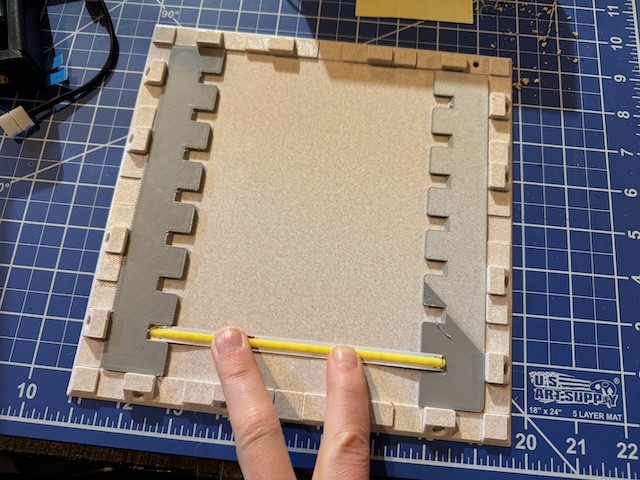 a hand holding down an LED strip between two crenelated pieces of plastic that are lying within a square of plastic that itself has crenelated edges.