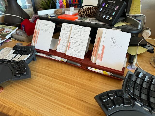 3 red plastic card holders with many offset slots each filled with one or more cards. In the front of each is a pen holder with a pen in it. below them you can see my keyboard halves and the clips holding them to the desk.