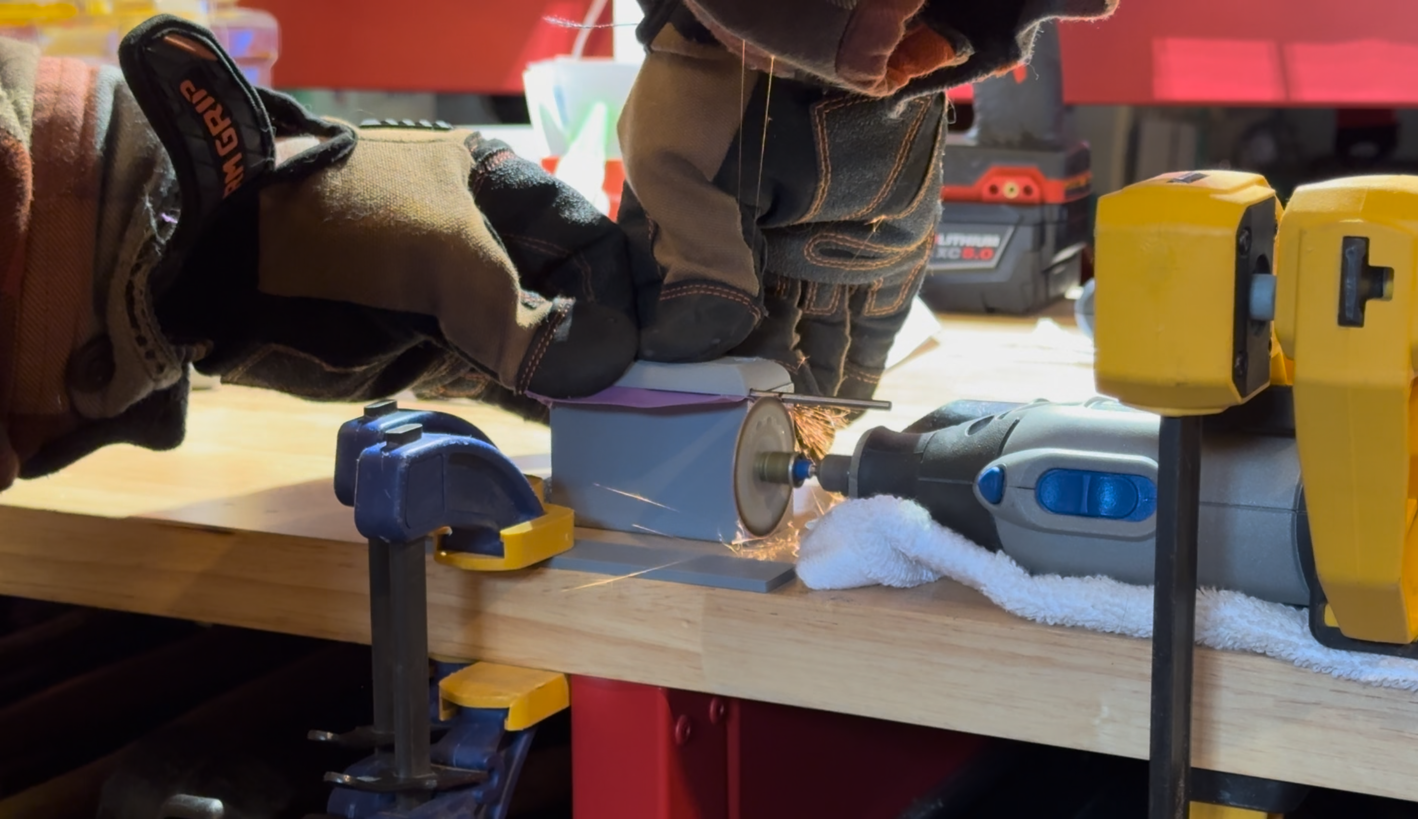 two gloved hands press against a grey brick with a rounded top and a thin purple sheet coming out of it. there's a steel rod coming out the side that is being pressed against a spinning Dremel blade from which sparks are shooting.